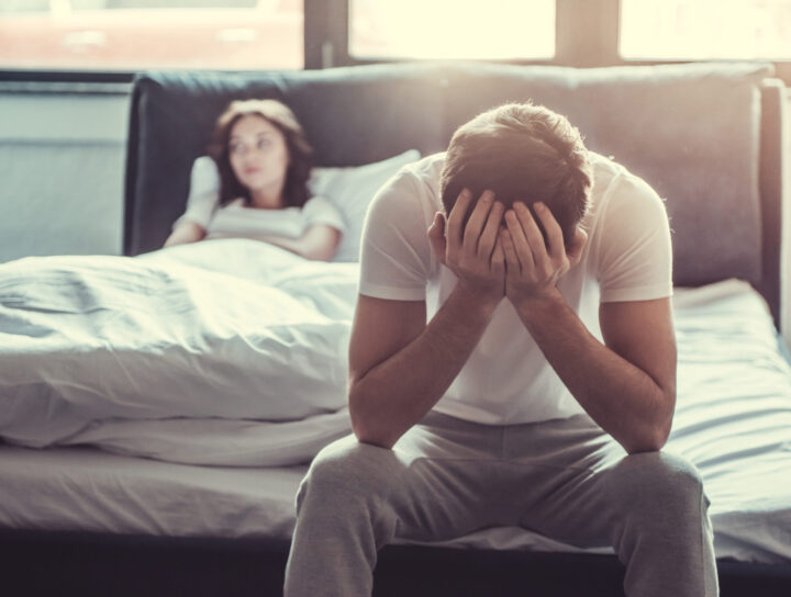 We have a problem. Upset young man sitting on the edge of the bed and holding his head against the background of a girlfriend, lying in bed.