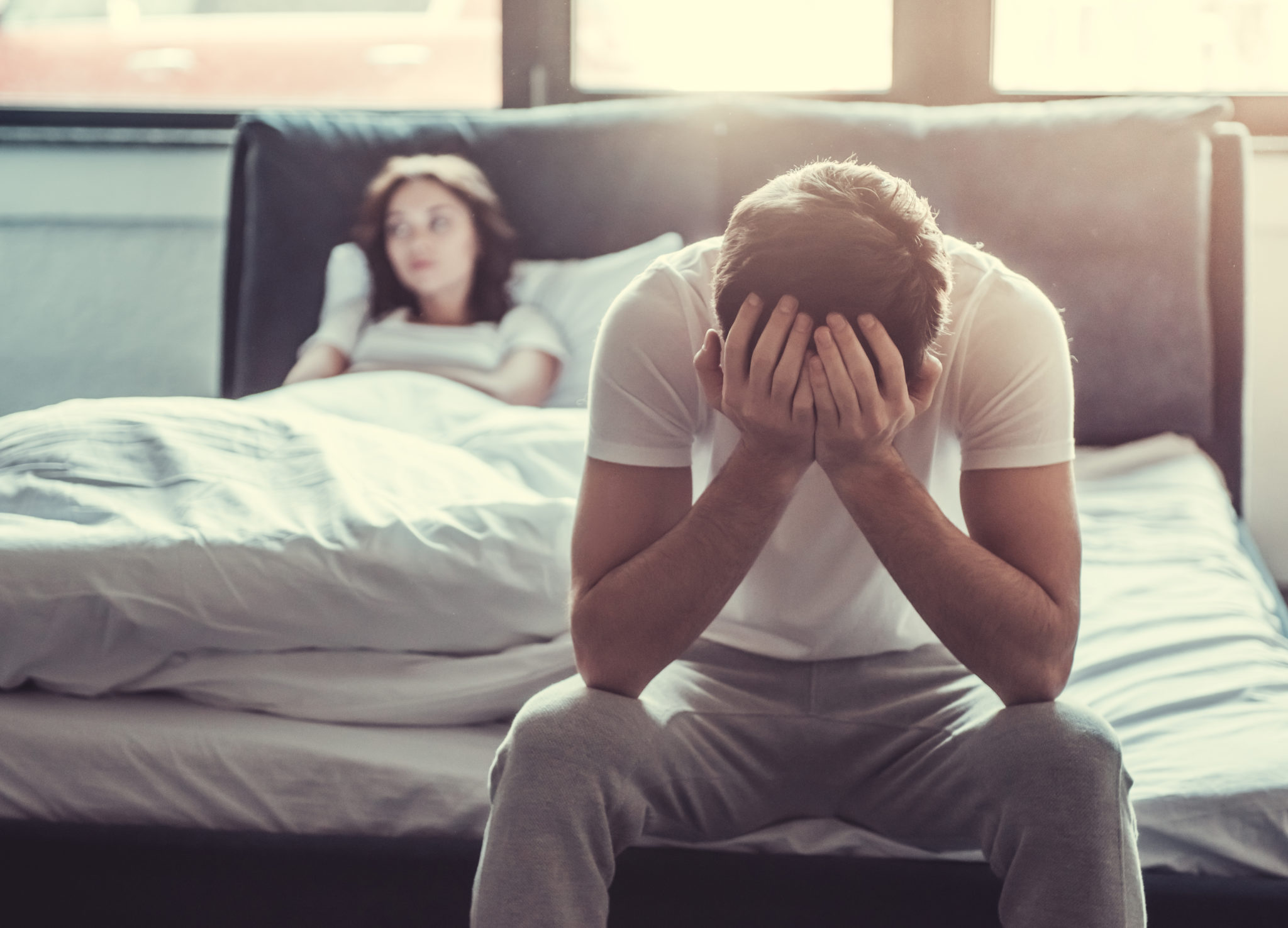 We have a problem. Upset young man sitting on the edge of the bed and holding his head against the background of a girlfriend, lying in bed.